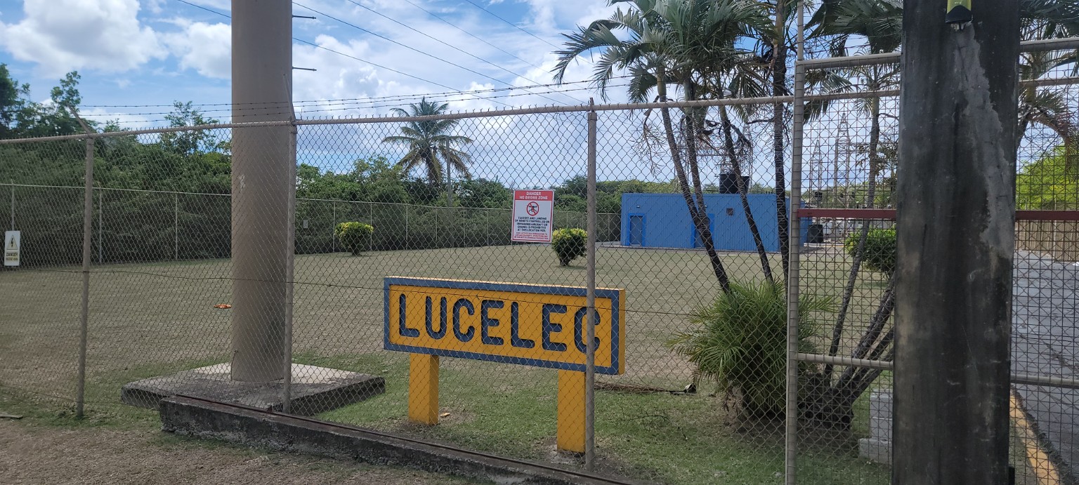 LUCELEC sign behind a chain-link fence at an electrical facility, with palms and utility structures nearby.