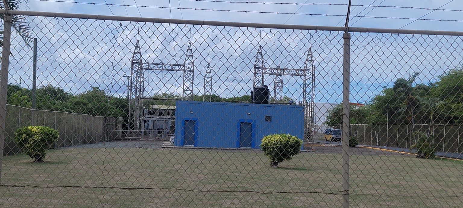 A blue utility building and electrical equipment seen through a chain-link perimeter fence.