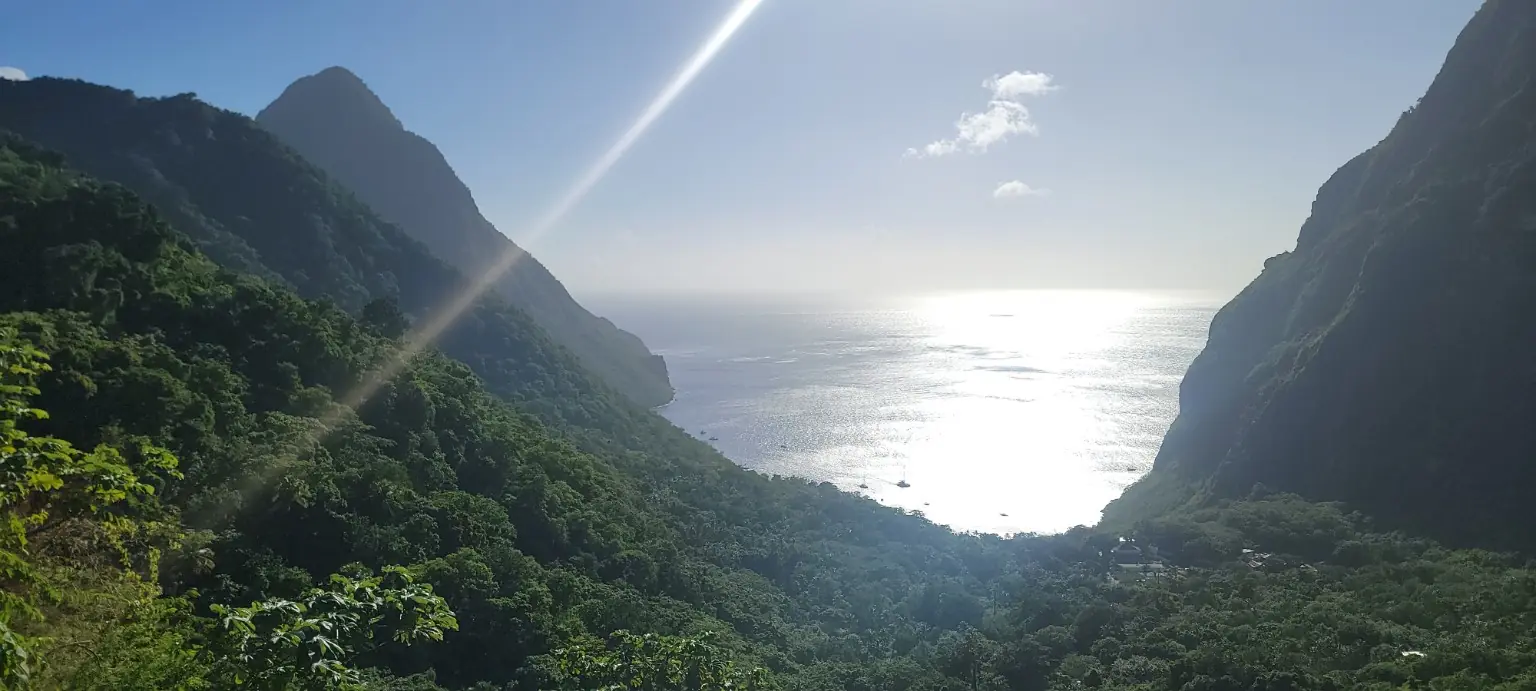 Forested Piton slopes frame a valley opening onto the sunlit sea, with small boats near the shore.