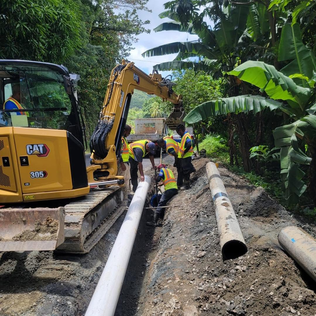 A crew lays a large water pipe beside an excavator on a narrow road
